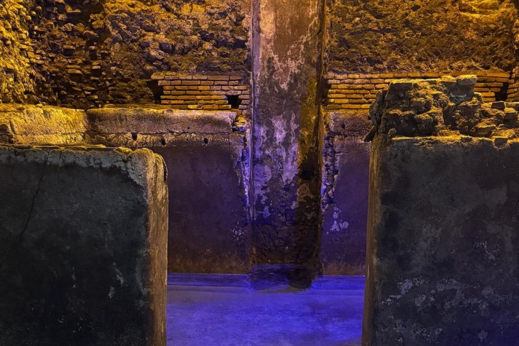 중앙 벽과 물이 가득한 전경이 특징인 노란색과 파란색 조명으로 빛나는 고대 석조 건축물입니다.