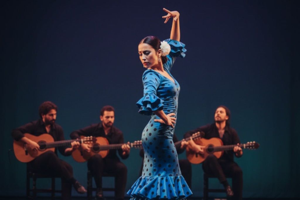 파란 물방울 무늬의 드레스를 입은 여성이 무대에서 플라멩코 춤을 추고 있으며, 4명의 기타리스트가 앉아 있습니다.