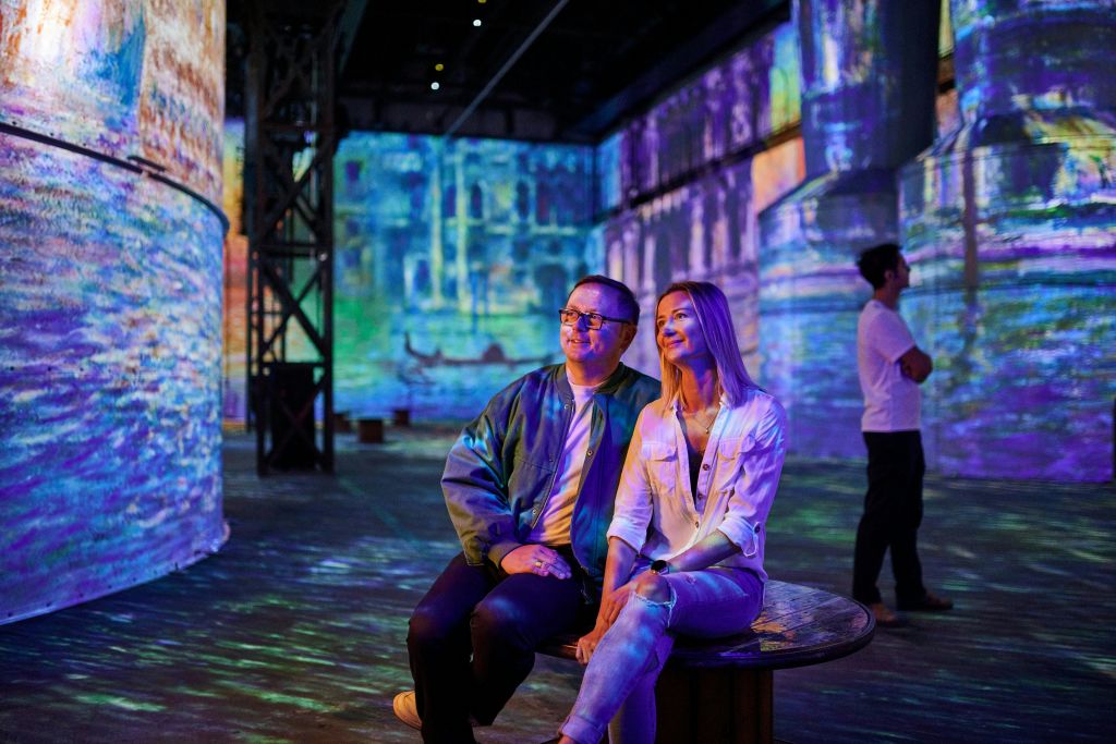 한 남녀가 벤치에 앉아 벽과 기둥에 화려하고 투영된 이미지로 둘러싸인 몰입형 미술 전시회를 관람하고 있습니다.