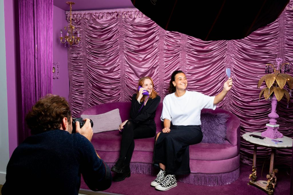 보라색 소파에 앉아 있는 두 명의 여성이 핸드헬드 거울을 들고 있습니다. 보라색으로 장식된 벽과 인테리어가 돋보이는 방에서 말이죠.