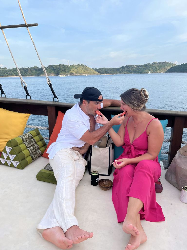 흰색 옷을 입은 남성과 분홍색 드레스를 입은 여성이 아름다운 물과 섬을 배경으로 보트에서 수박 조각을 먹고 있습니다.