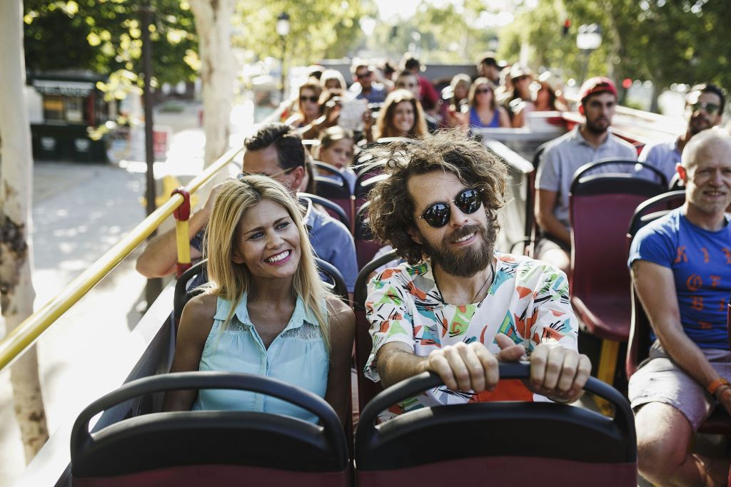 2층 투어 버스에 앉은 사람들은 화창한 날 웃으며 라이딩을 즐깁니다. 나무와 가로등이 보입니다.