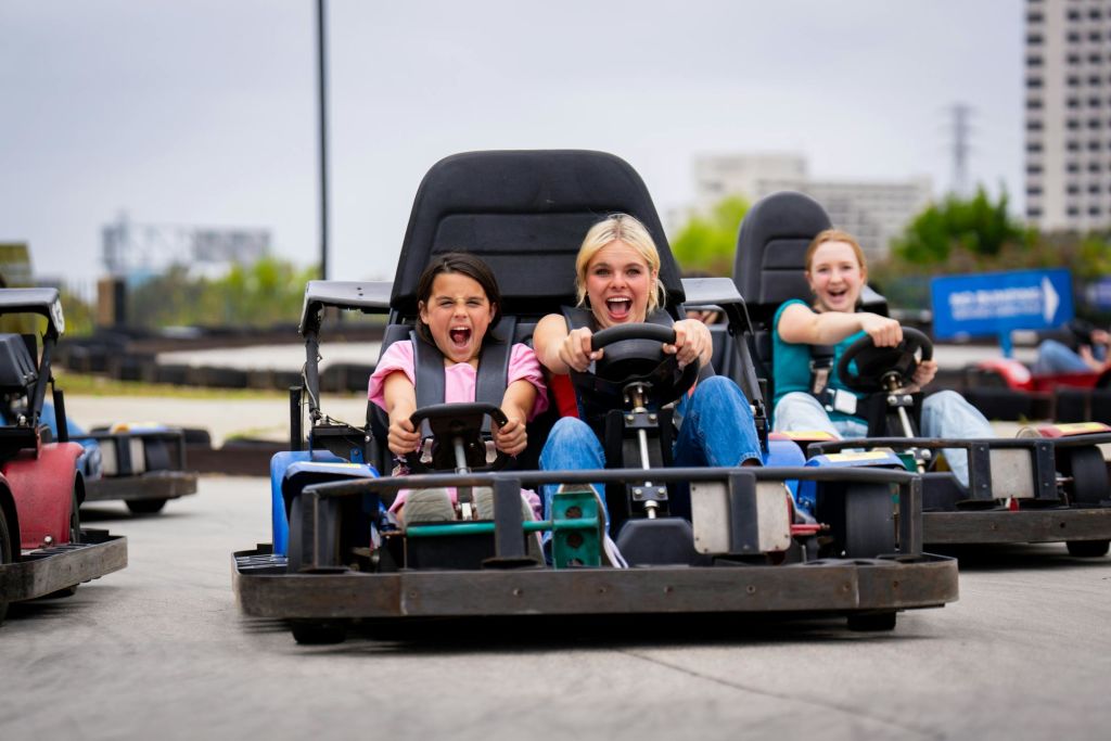 트랙에서 고카트를 타고 열정적으로 달리는 두 사람, 한 사람은 스티어링을 하고 다른 한 사람은 응원하며, 세 번째 사람은 부분적으로 보입니다.