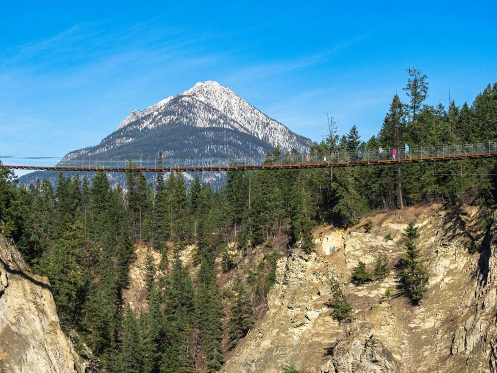 숲이 우거진 골짜기 너머로 높이 솟은 현수교를 걷고 있는 사람들로, 눈 덮인 산을 배경으로 걸어가고 있습니다.