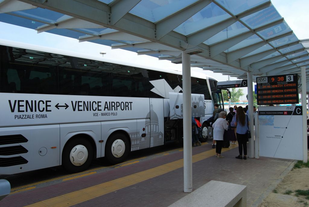 지붕이 있는 터미널에서 '베네치아 공항' 이라고 적힌 버스에 탑승하는 사람들. 근처 표지판에는 "C" 와 버스 목적지가 표시됩니다.