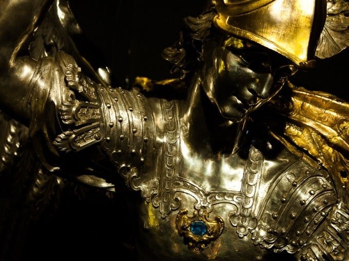 파란색 원석과 날개 달린 어깨 패드가 달린 디테일한 갑옷을 입은 전사를 묘사한 금속 조각상을 클로즈업했습니다.