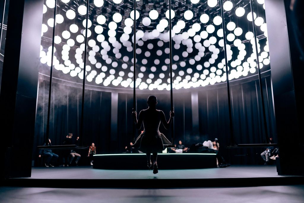 흰색 불빛이 반짝이는 원형 천장 아래 그네를 타는 사람으로, 방을 배경으로 사람들이 둘러앉아 있습니다.