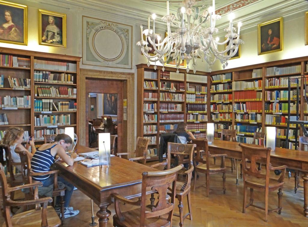 벽에 놓인 책장, 샹들리에, 액자에 담긴 초상화로 둘러싸인 서재의 나무 테이블에서 공부하는 사람들.