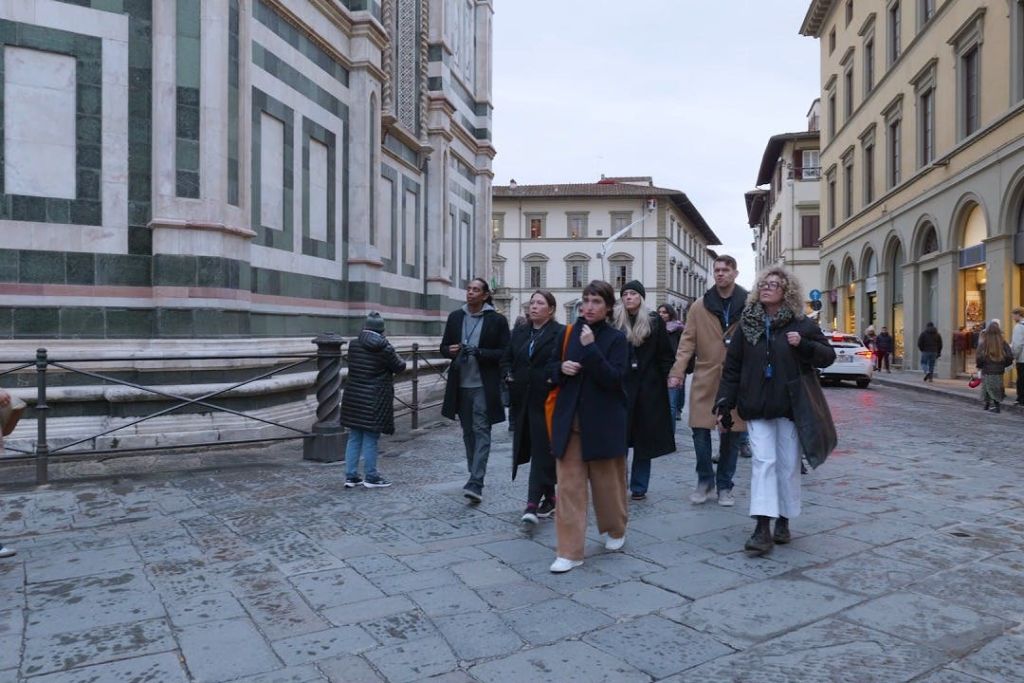 유럽 도시의 유서 깊은 건물 옆 자갈길을 함께 걷는 한 무리의 사람들.