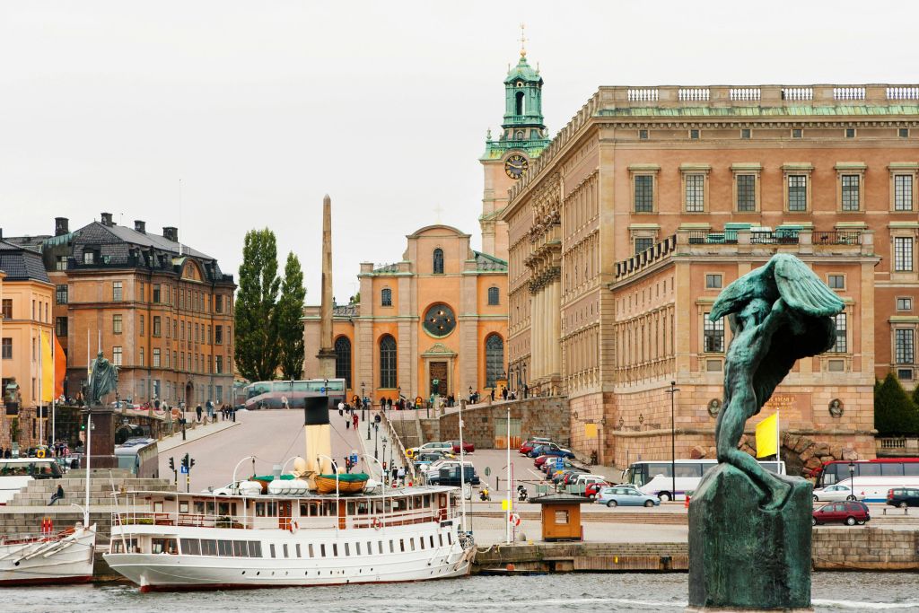 시계탑이 있는 교회와 근처를 걷는 사람들 등 유서 깊은 건물이 있는 해안가에 정박한 흰색 보트입니다.