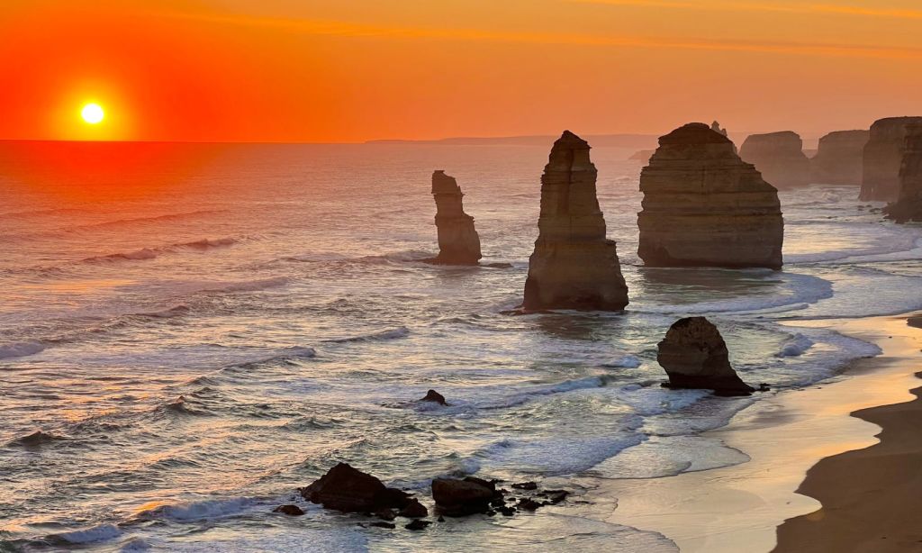 높은 암석 지층이 바다에서 활기찬 오렌지빛 석양을 배경으로 솟아오르고, 해안에는 파도가 부드럽게 부서집니다.