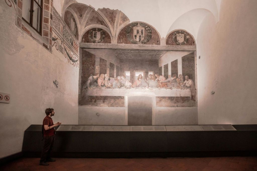 한 사람이 조명이 희미한 갤러리의 레오나르도 다빈치 (Leonardo da Vinci) 의 "최후의 만찬 (The Last Supper)" 벽화 앞에 서 있습니다.