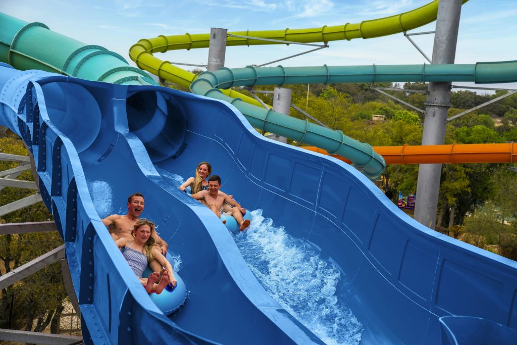 푸른 물 미끄럼틀을 타는 두 쌍의 사람들이 웃으며 튀고, 녹색과 노란색 미끄럼틀을 배경으로 합니다.