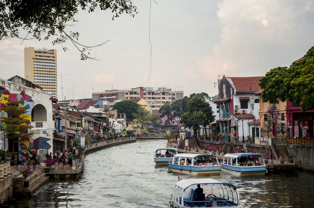 몇몇 투어 보트는 나무와 건물이 있는 도시 지역을 통해 강을 항해하고 사람들은 강변 산책로를 따라 걷습니다.