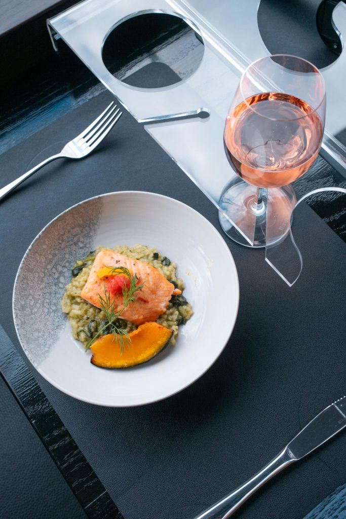 식탁에 놓인 로제 와인, 포크, 플레이스매트 옆에는 연어와 리조또 한 접시에 허브가 장식되어 있습니다.