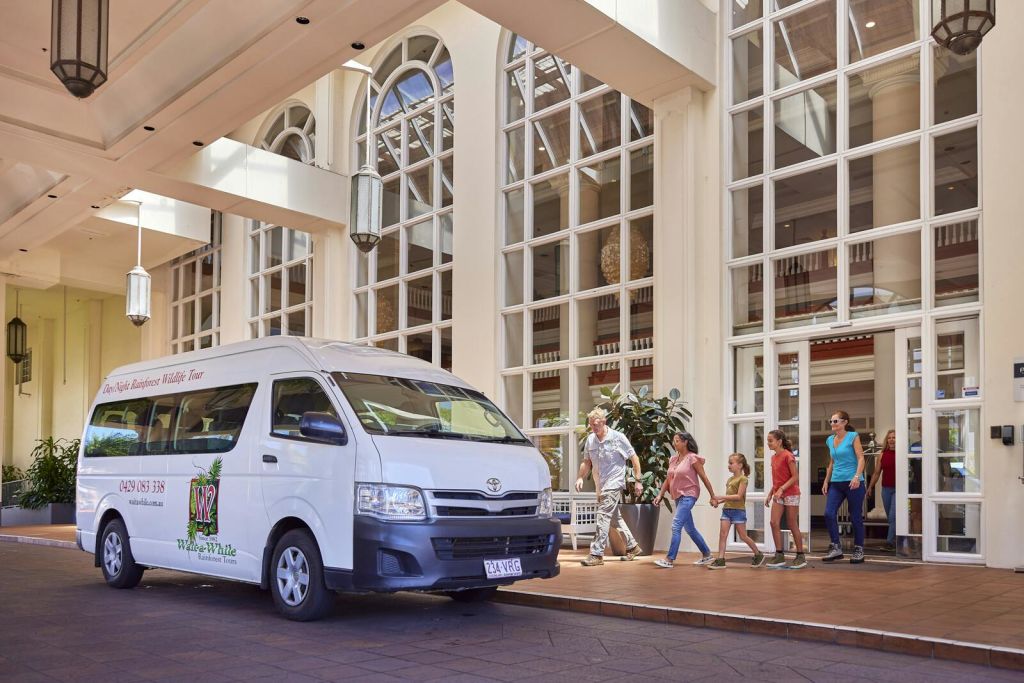 커다란 창문이 있는 건물 앞에 흰색 밴이 주차되어 있으며, 어른과 어린이를 포함한 일행이 지나갑니다.