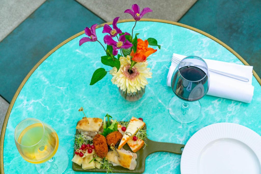 청록색 테이블에 다채로운 식사가 준비되어 있으며, 치즈 플래터, 레드 와인, 꽃무늬 센터피스가 꽃병에 담겨 있습니다.