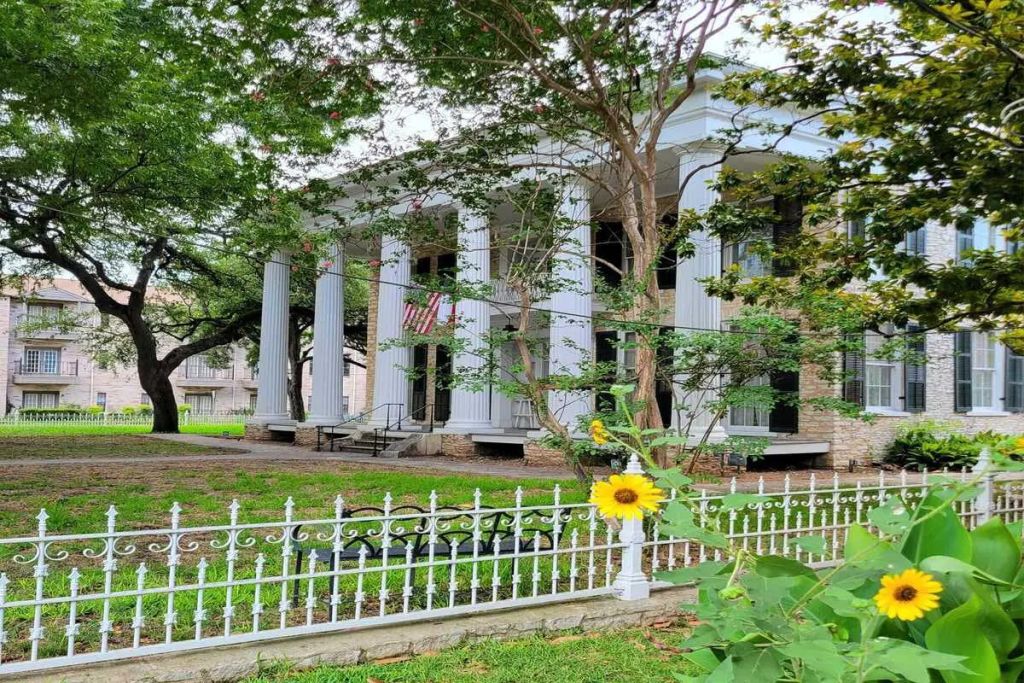 나무로 둘러싸인 높은 흰색 기둥과 미국 국기가 있는 역사적인 건물로, 앞에 해바라기가 있는 흰색 연철 울타리가 있습니다.