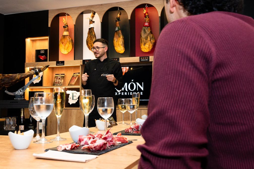 검은색 셰프의 코트를 입은 한 남성이 경화 햄, 와인잔, 음식 접시를 들고 요리를 즐기는 청중에게 말을 걸고 있습니다.