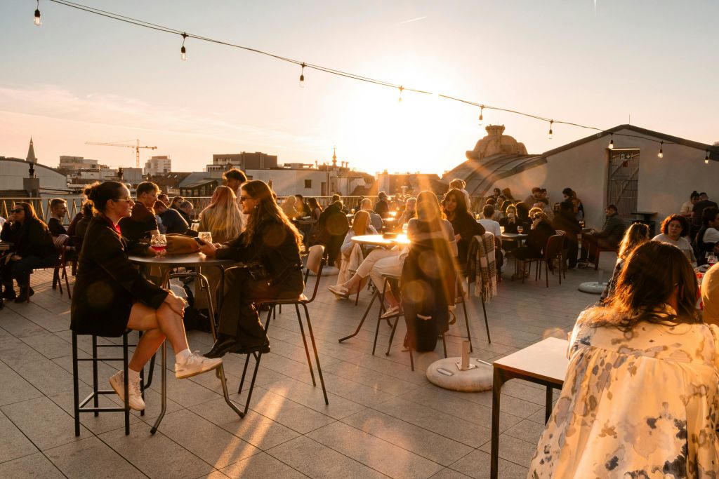 해 질 녘 옥상 파티오의 테이블에 앉아 있는 사람들로, 도시 건물을 배경으로 머리 위로 조명이 켜져 있습니다.