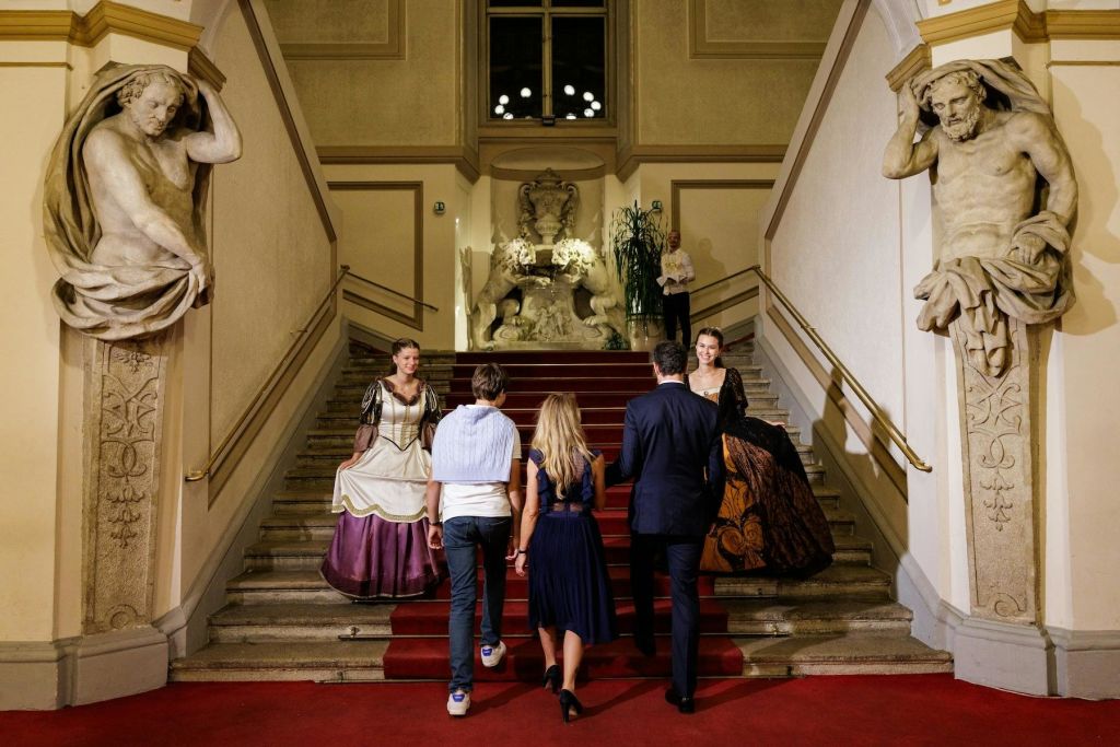 격식을 차린 사람들이 레드 카펫을 깔고 웅장한 계단을 올라가고, 우아한 인테리어에 클래식한 조각상과 화려한 장식이 둘러싸여 있습니다.