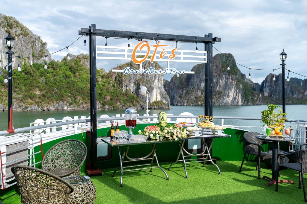 야외 데크에는 의자, 음료, 음식이 놓인 테이블, 경치를 배경으로 한 '오티스의 크루즈 투어 & 여행' 표지판이 설치되어 있습니다.