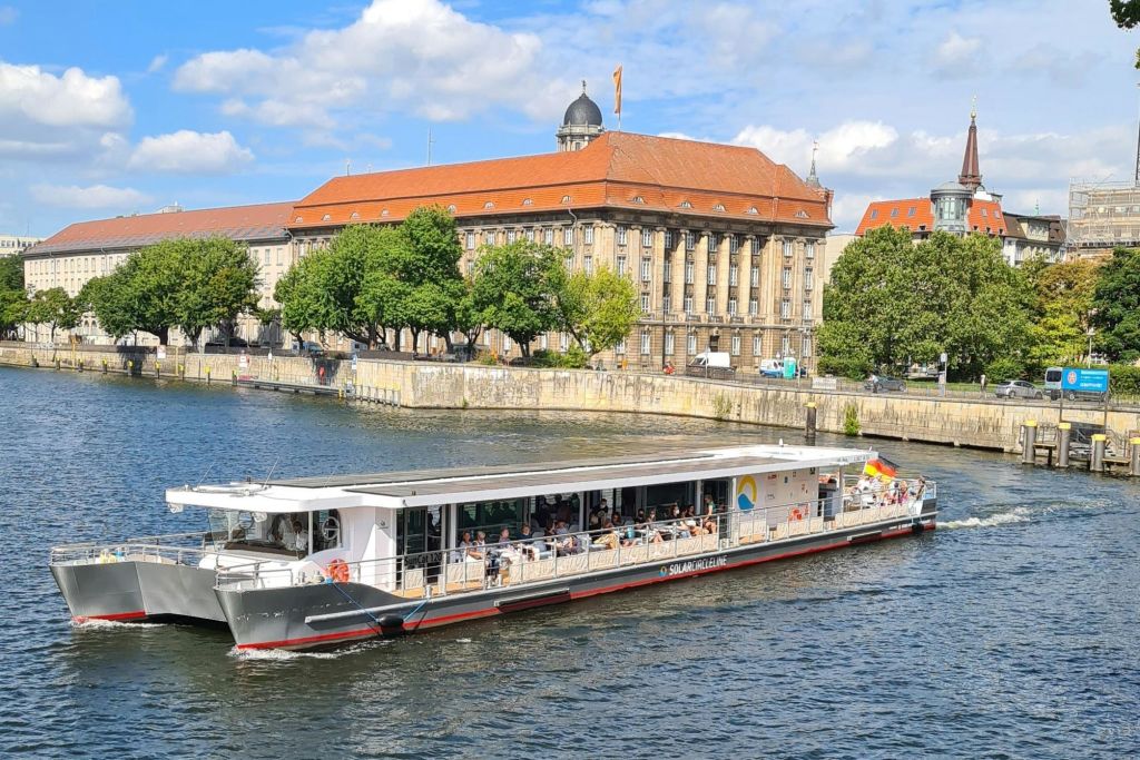 사람들로 가득 찬 강이 흐르고 있습니다. 배경에는 붉은 지붕과 나무가 있는 유서 깊은 건물이 있습니다.