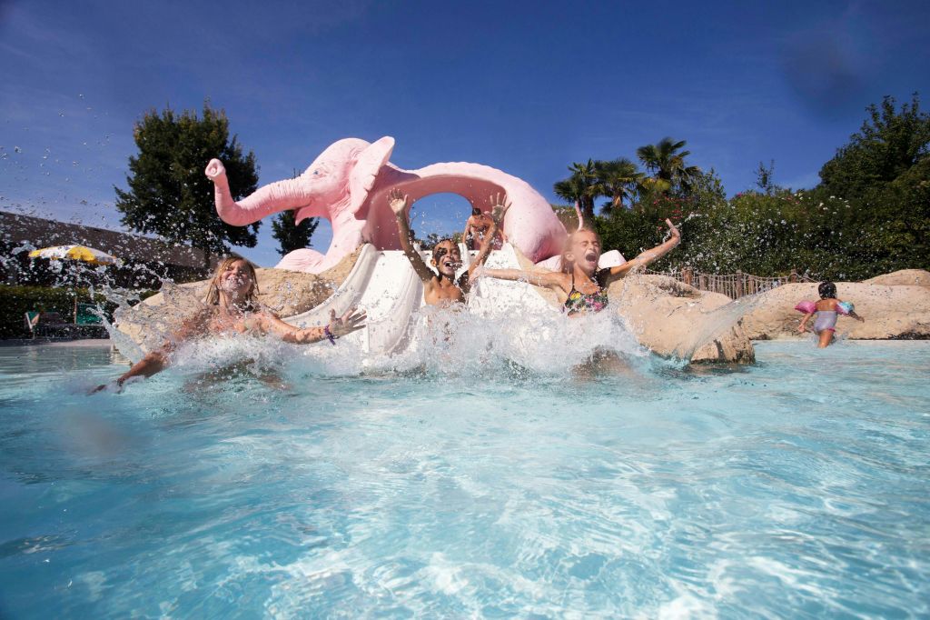 분홍색 코끼리 모양의 미끄럼틀에서 수영장에 미끄러져 들어가 맑고 푸른 하늘 아래에서 물이 튀고 있습니다.