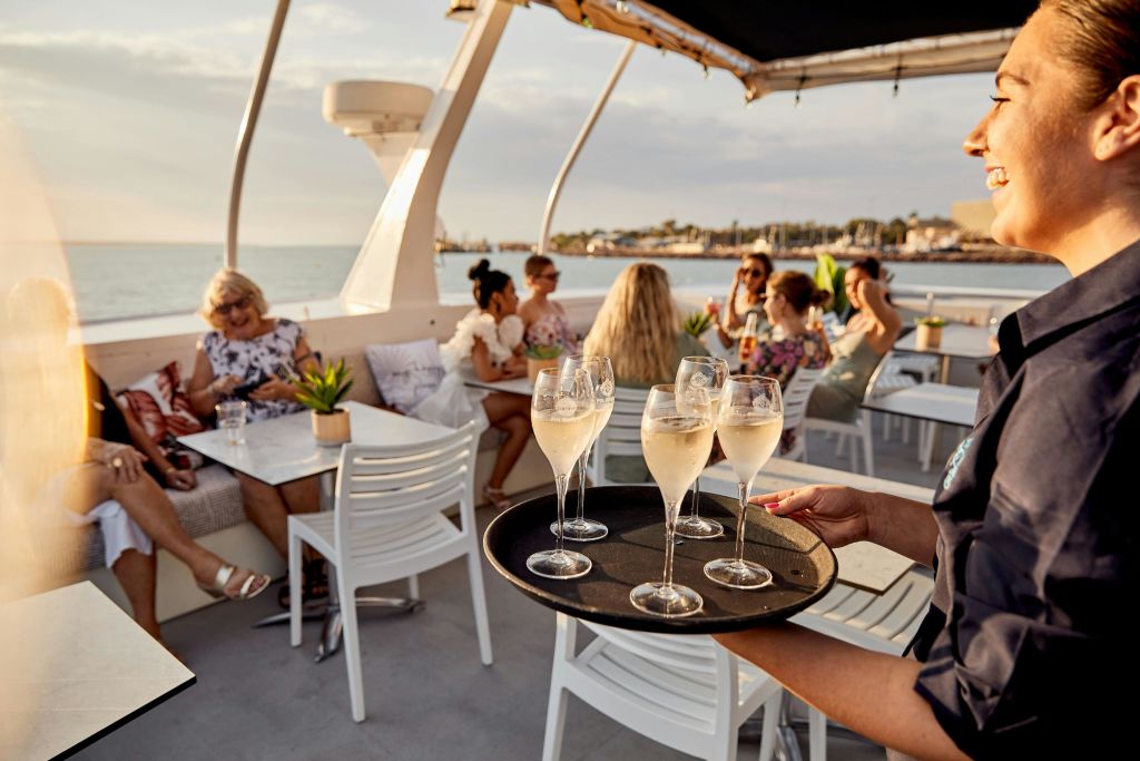 웃고 있는 웨이트리스가 보트 갑판에 샴페인 잔을 쟁반에 올려놓고 사람들과 어울리고 있습니다.