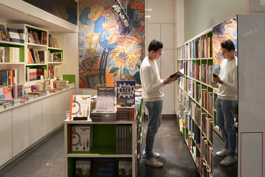 흰색 스웨터와 파란색 청바지를 입은 남성이 서점에서 화려한 벽걸이 예술품과 책으로 가득 찬 선반이 있는 책을 읽고 있습니다.