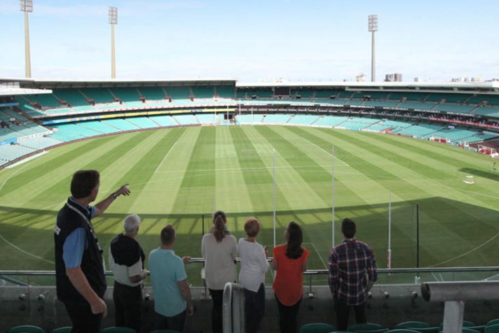 한 무리의 사람들이 경기장에 서서 맑은 하늘 아래 골 포스트가 있는 넓은 녹색 필드를 보고 있습니다.