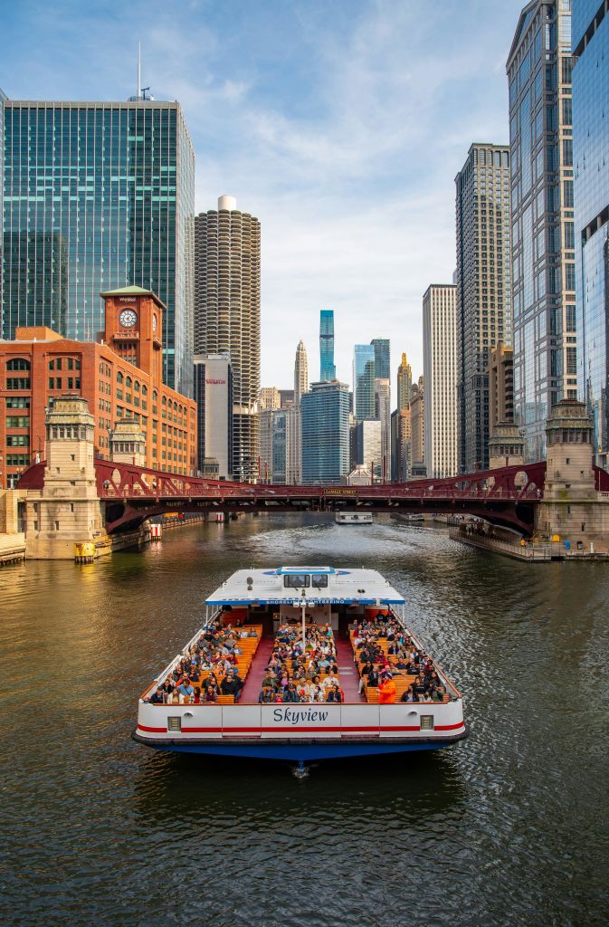 높은 고층 빌딩과 현대적인 건축물이 있는 도시의 붉은 다리에 사람들이 가득한 보트가 다가옵니다.