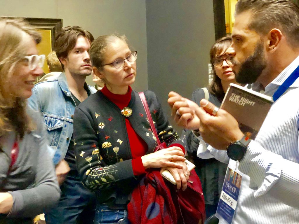 꽃무늬 스웨터에 안경을 쓴 여성을 포함한 한 무리의 사람들은 손으로 제스처를 취하는 사람의 말을 주의 깊게 듣습니다.