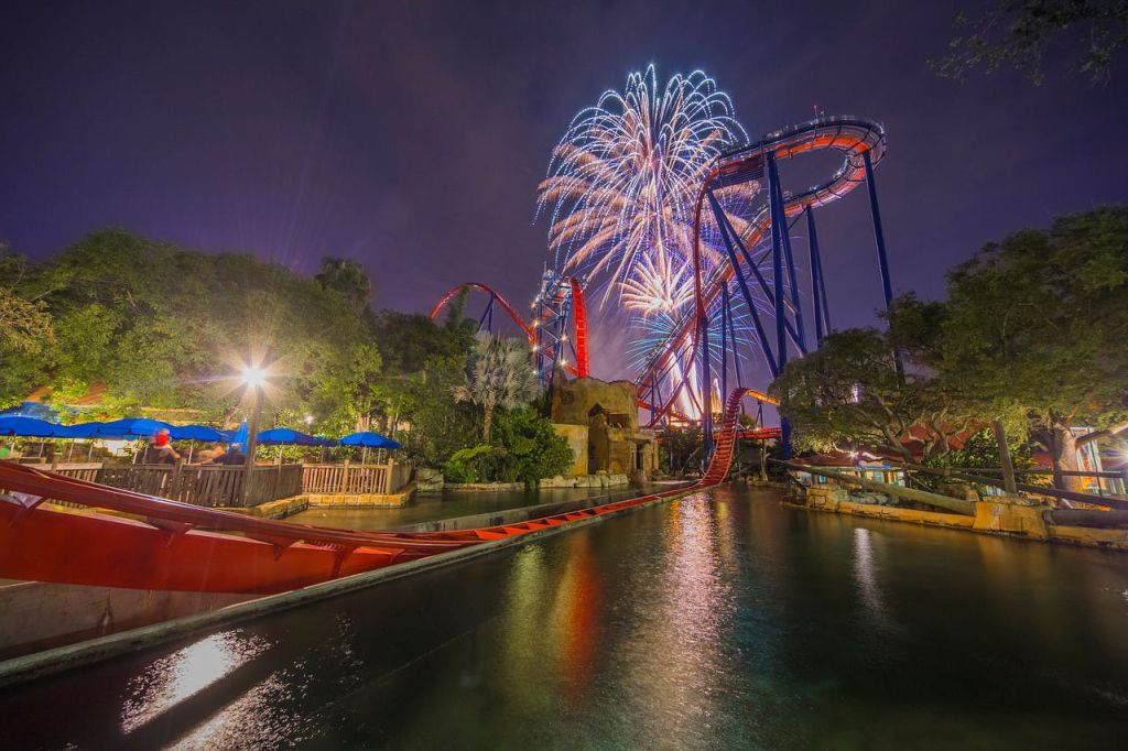 불꽃놀이는 나무와 구조물로 둘러싸인 물에 반사되는 트랙과 함께 밤에 밝은 조명으로 빛나는 롤러코스터 위에서 폭발합니다.