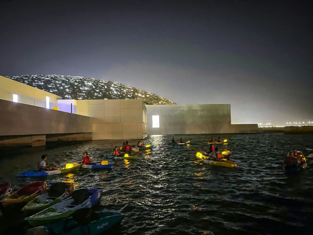 격자 무늬의 돔 모양 지붕을 배경으로 현대적인 건물 근처의 어둡고 밝은 수변에서 카약을 타는 사람들.