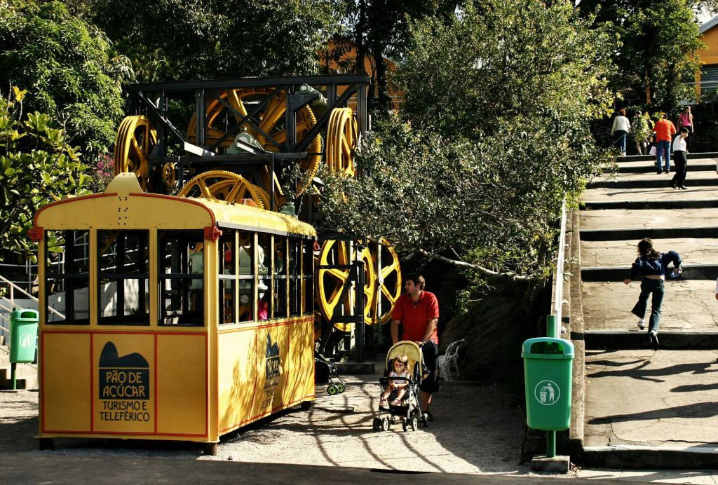 한 남성이 노란색 트램과 녹색 쓰레기통 근처에서 유모차를 밀고 있으며, 배경에는 커다란 노란색 기어가 있고 머리 위에는 나무가 있습니다.