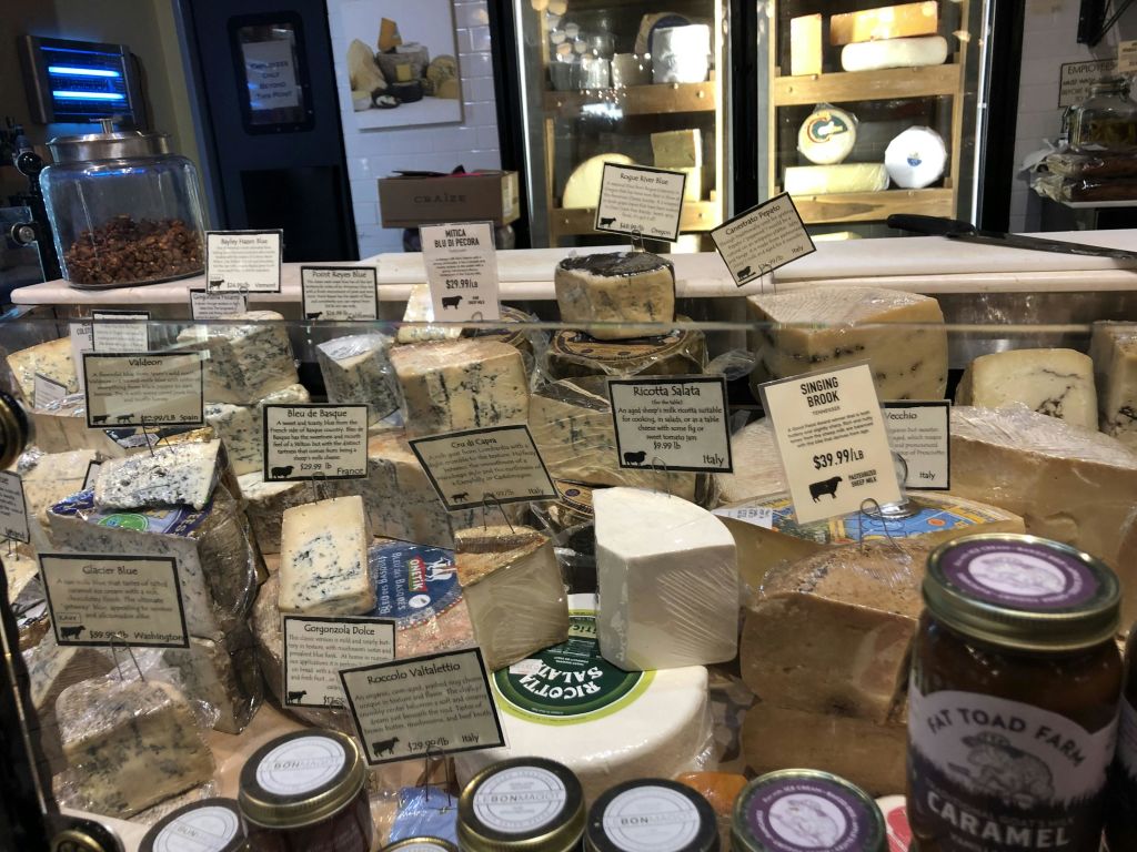 전경에 조미료 항아리가 있는 델리 세팅에 블루 치즈와 고우다를 포함한 다양한 치즈 라벨이 표시되어 있습니다.
