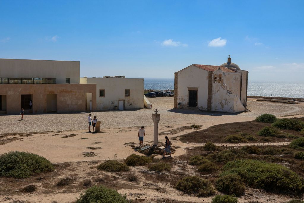 해안선 근처에 있는 작은 예배당과 인접한 건물로, 사람들이 맑고 푸른 하늘 아래에서 걷고 서 있습니다.