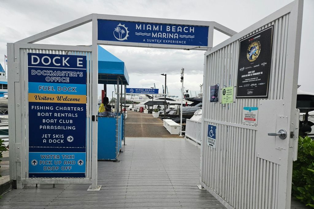 다양한 서비스를 받을 수 있는 표지판이 있는 마이애미 비치 마리나 입구 게이트, 배경에 정박한 보트, 머리 위로 흐린 하늘.