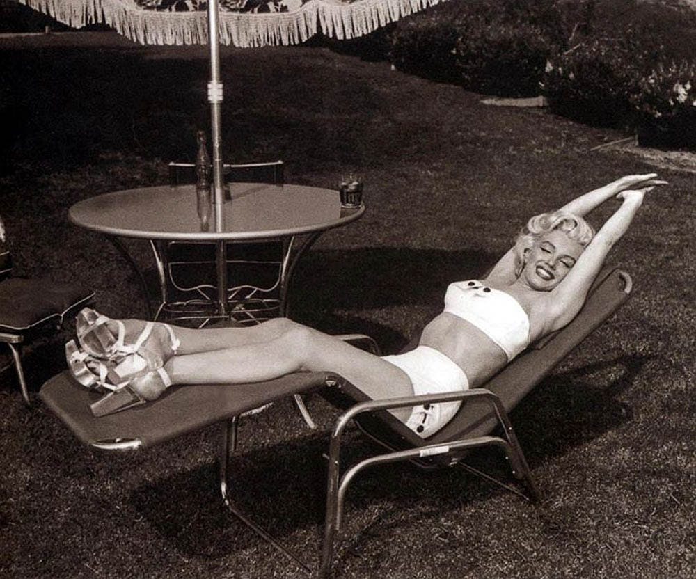 투피스 수영복을 입은 여성이 프린지 우산이 달린 원형 테이블 옆 선베드에서 팔을 머리 위로 뻗은 채 웃고 있습니다.