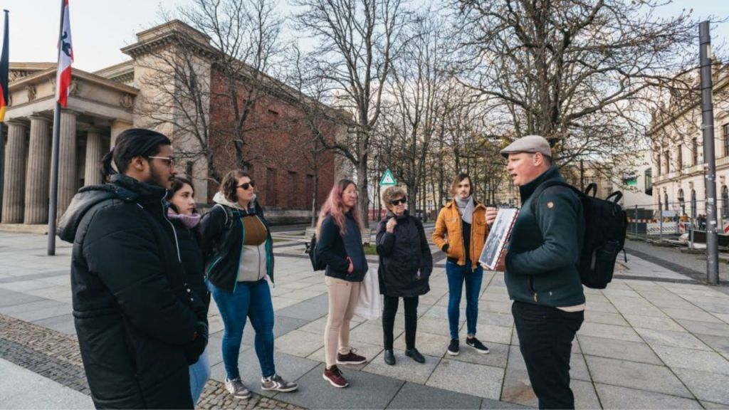 흐린 날 깃대와 잎이 없는 나무가 있는 역사적인 건물 근처에 한 무리의 사람들이 서 있습니다.