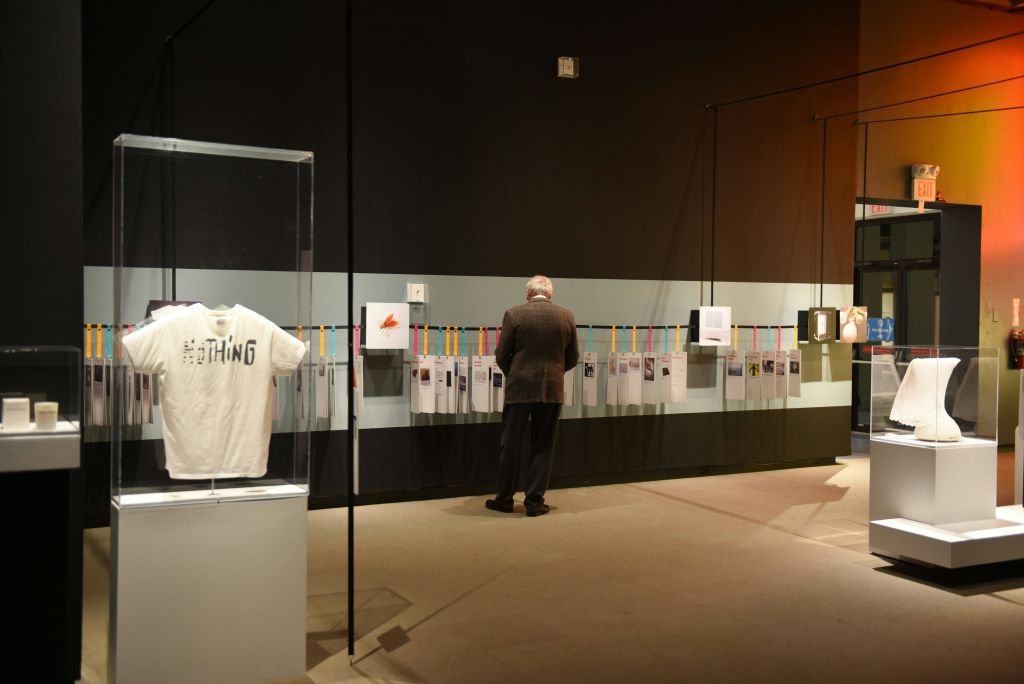 박물관에 있는 한 노인이 벽에 걸린 서류를 살펴보고 있습니다. 전면에 티셔츠가 달린 유리 케이스가 보입니다.