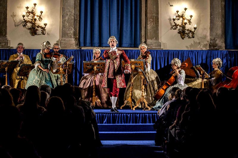 18세기 복장을 한 연주자들은 화려한 음악 스탠드와 샹들리에가 있는 파란색 무대에서 현악기를 부르고 연주합니다.