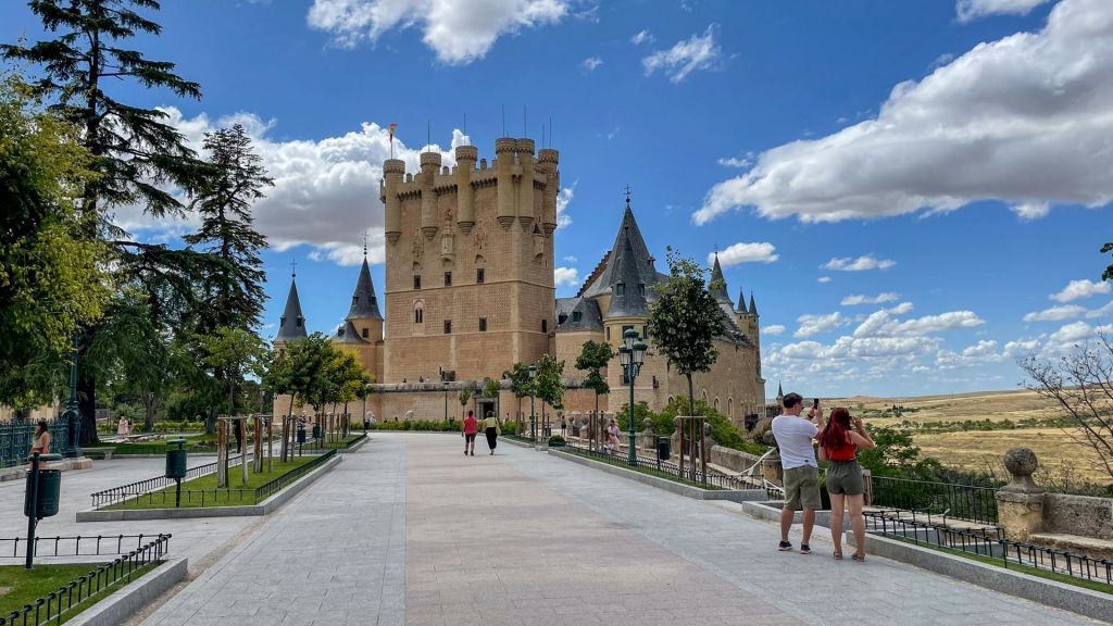 푸른 하늘 아래 높이 솟은 탑과 첨탑이 있는 웅장한 성으로 흩어져 있는 구름을 배경으로 관광객들이 사진을 찍고 있습니다.