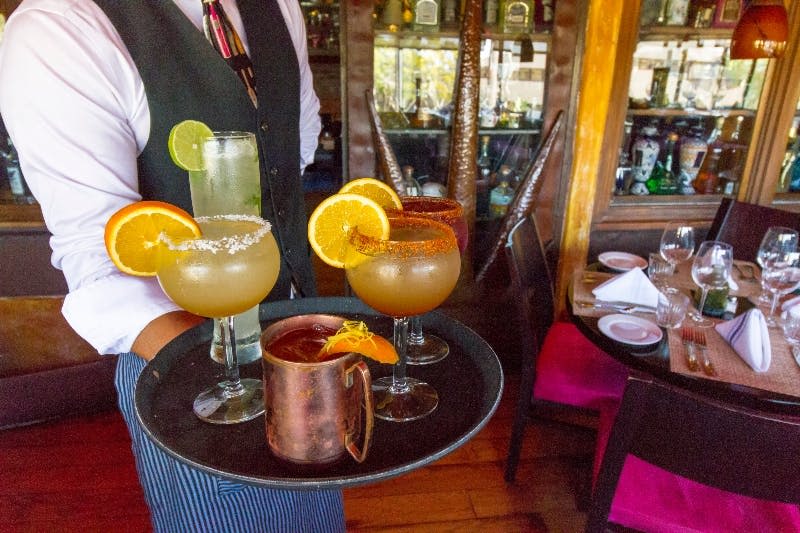 조끼와 넥타이를 입은 웨이터가 레스토랑에서 시트러스 슬라이스로 장식된 여러 칵테일이 담긴 트레이를 들고 있습니다.
