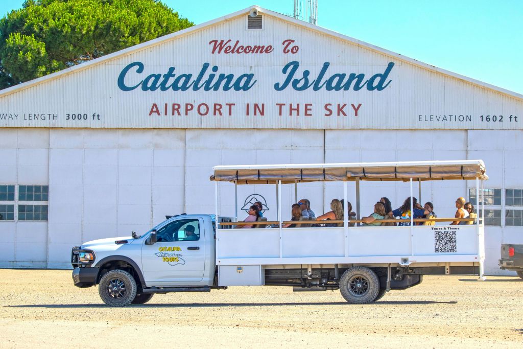 "하늘의 카탈리나 섬 공항에 오신 것을 환영합니다" 라는 표지판이 있는 건물 앞에 승객이 탑승한 투어 차량이 주차되어 있습니다.