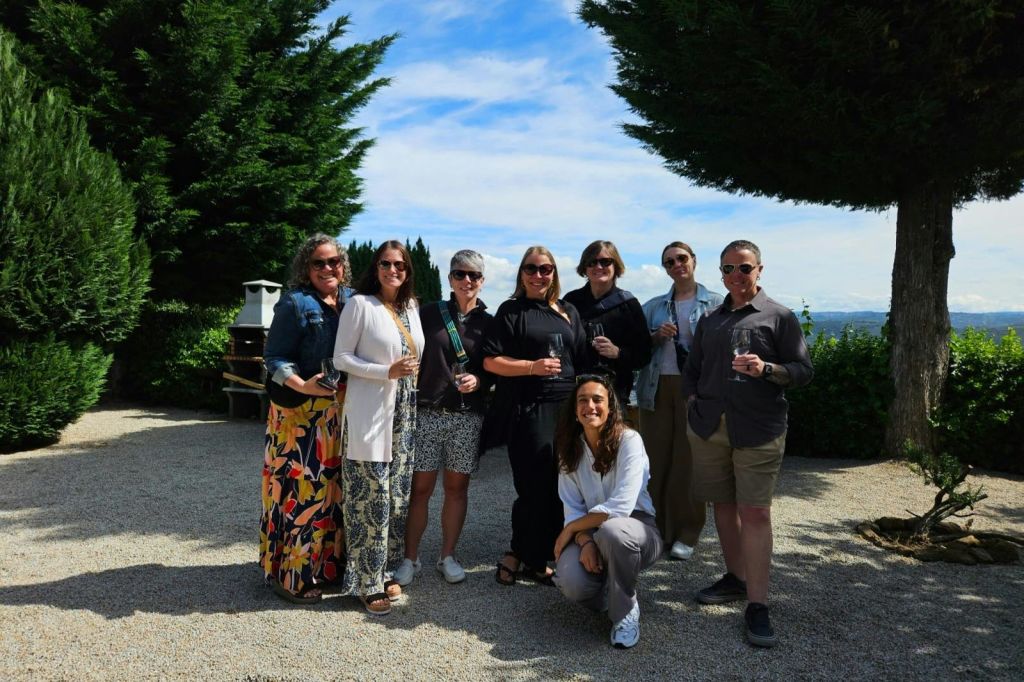 8명이 야외에 서서 미소를 짓고 음료를 들고 있습니다. 구름이 흩날리는 푸른 하늘 아래 녹음으로 둘러싸여 있습니다.