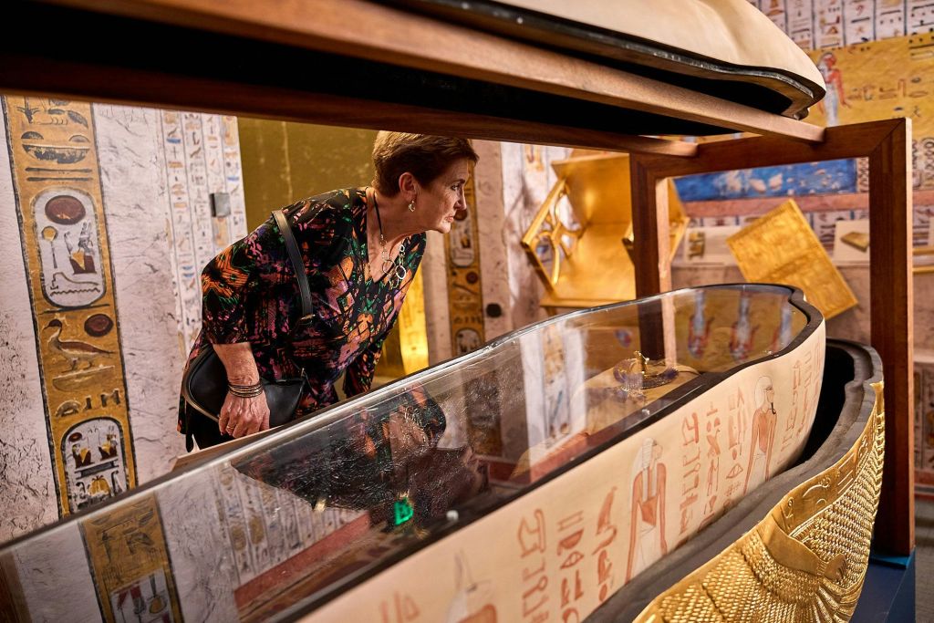한 여성이 박물관에 전시된 이집트 유물이 담긴 유리 진열 케이스를 들여다보고 있습니다. 벽에 상형 문자가 그려져 있습니다.