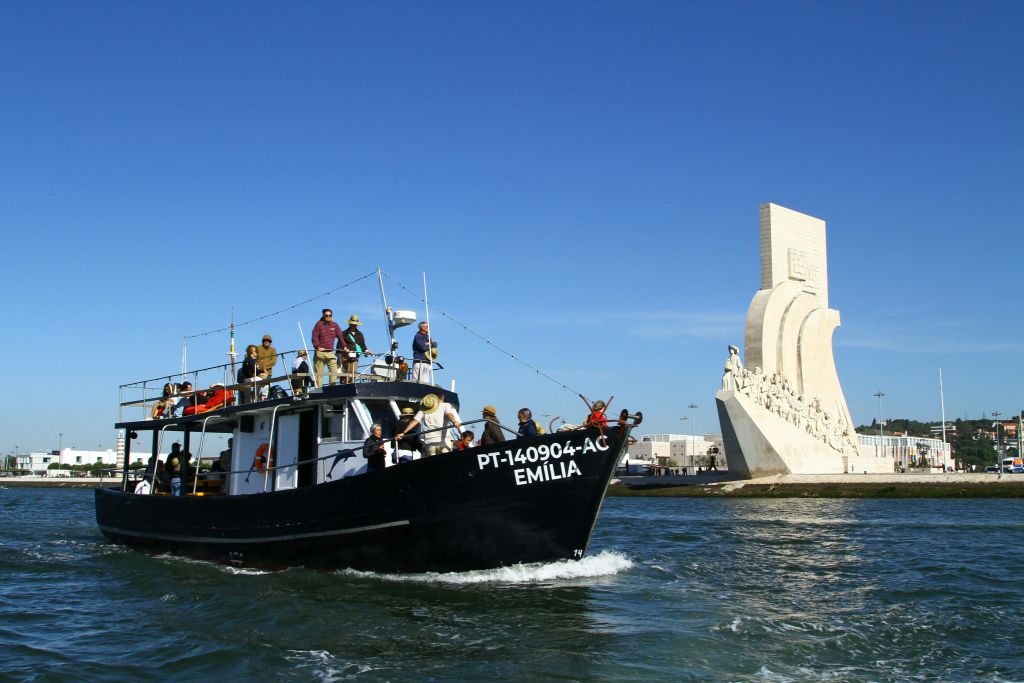 맑고 푸른 하늘 아래 커다란 해양 기념물 옆에 여러 명이 타고 있는 "에밀리아" 라는 이름의 검은 보트가 있습니다.