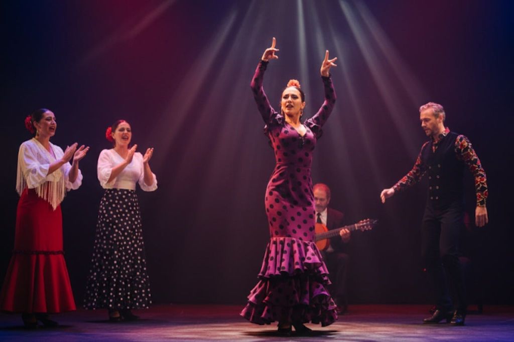 무대에서 퍼플 폴카 도트 드레스를 입은 플라멩코 댄서가 박수를 치는 개인과 기타리스트와 함께 공연합니다.